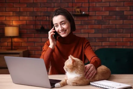 Uma mulher a falar ao telemóvel e a olhar para o portátil enquanto faz festas ao gato